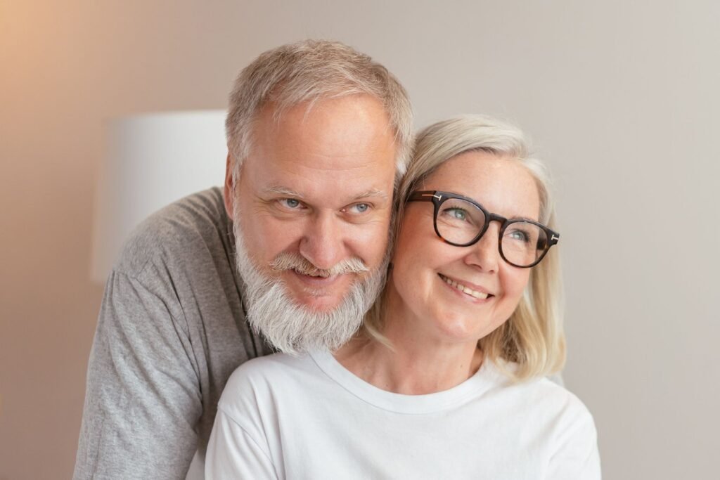 A smiling older couple embracing at home, the man with a gray beard behind the woman, who wears dark-rimmed glasses
