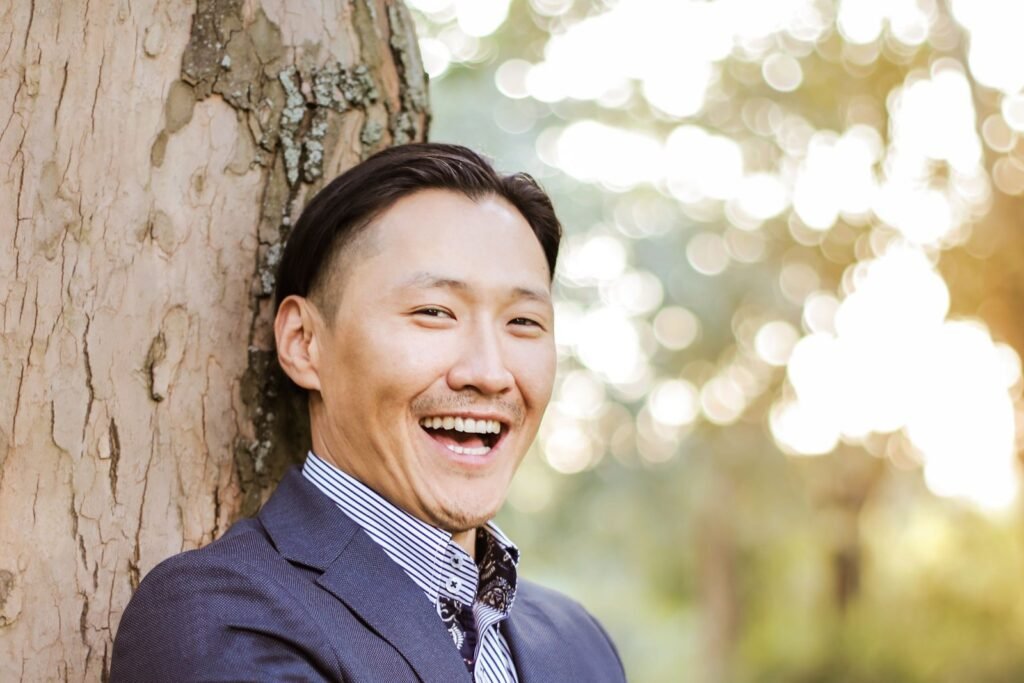 A young man in a navy blazer laughing with a wide smile as he leans against a tree in golden evening light